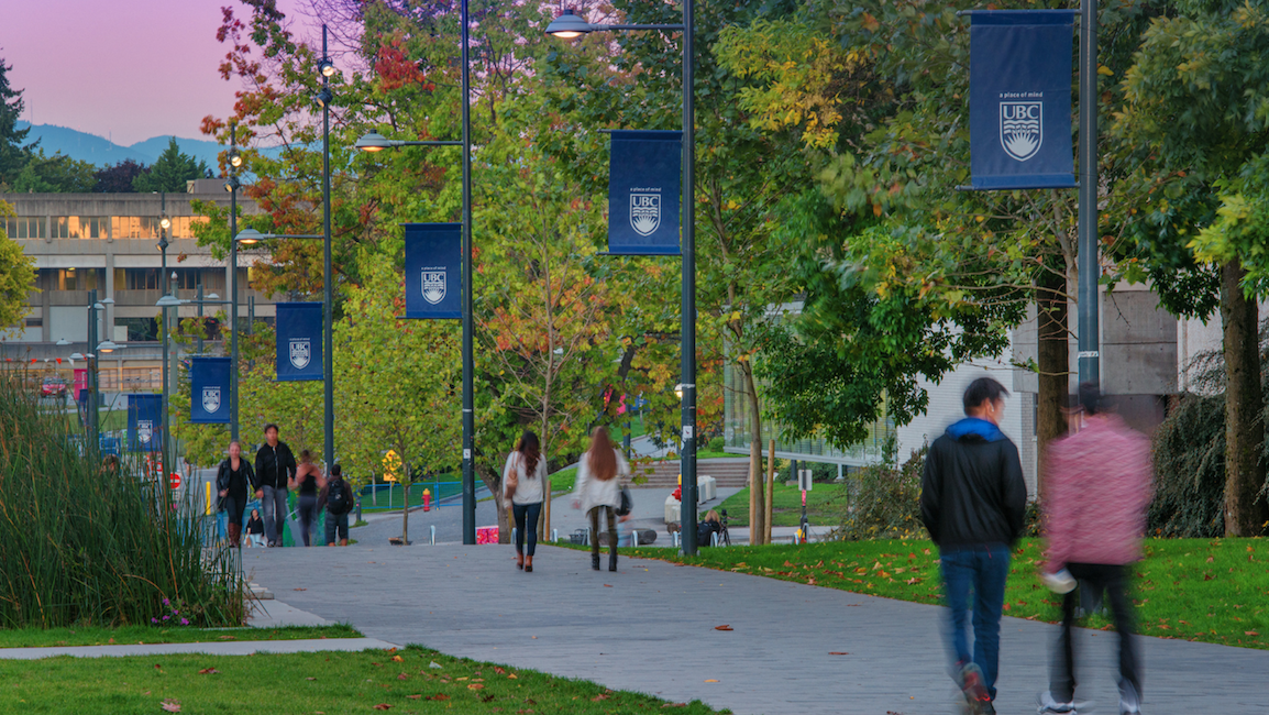 Image of the UBC campus at sunset with a walkway of students and trees and mountains in view. 