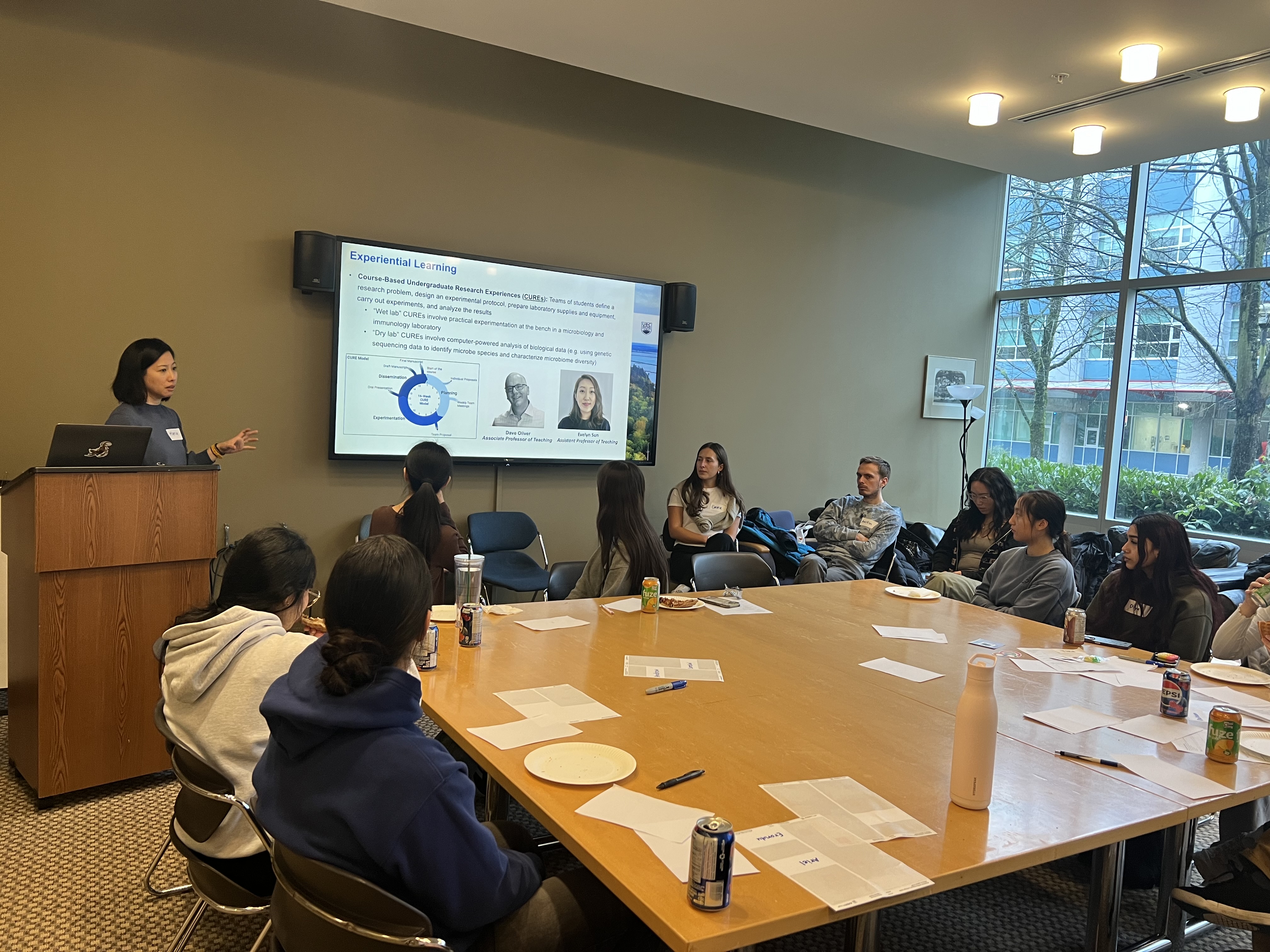 A person standing at a podium lectures to a group of people sitting around a table.