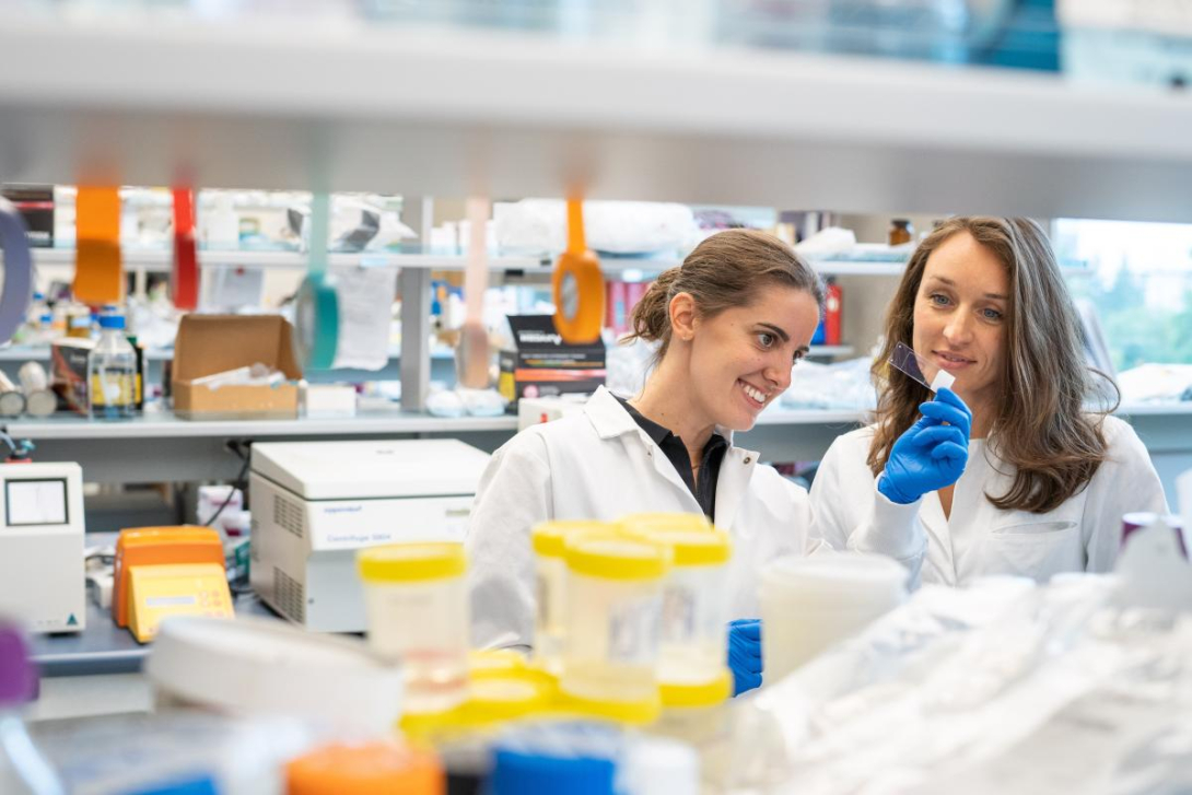 Sophie Cotton and Carolina Tropini wearing lab coats and looking at a sample in their lab.